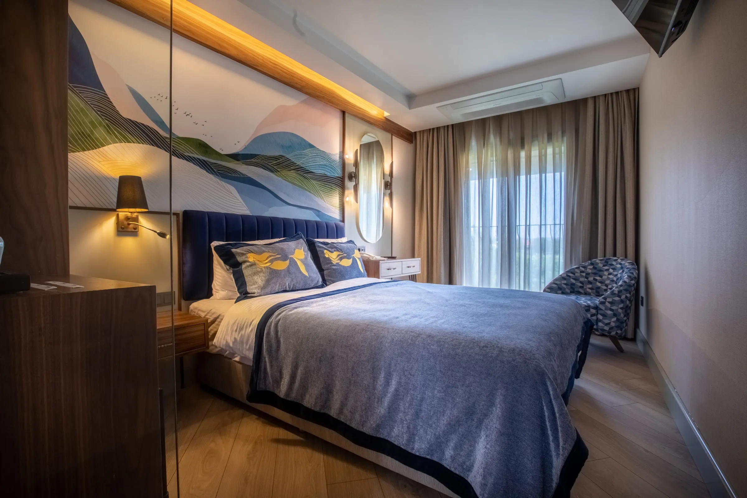 A bed with a petrol velvet headboard, cushions with a gold mermaid motif and a wave mural — one of the hotel rooms