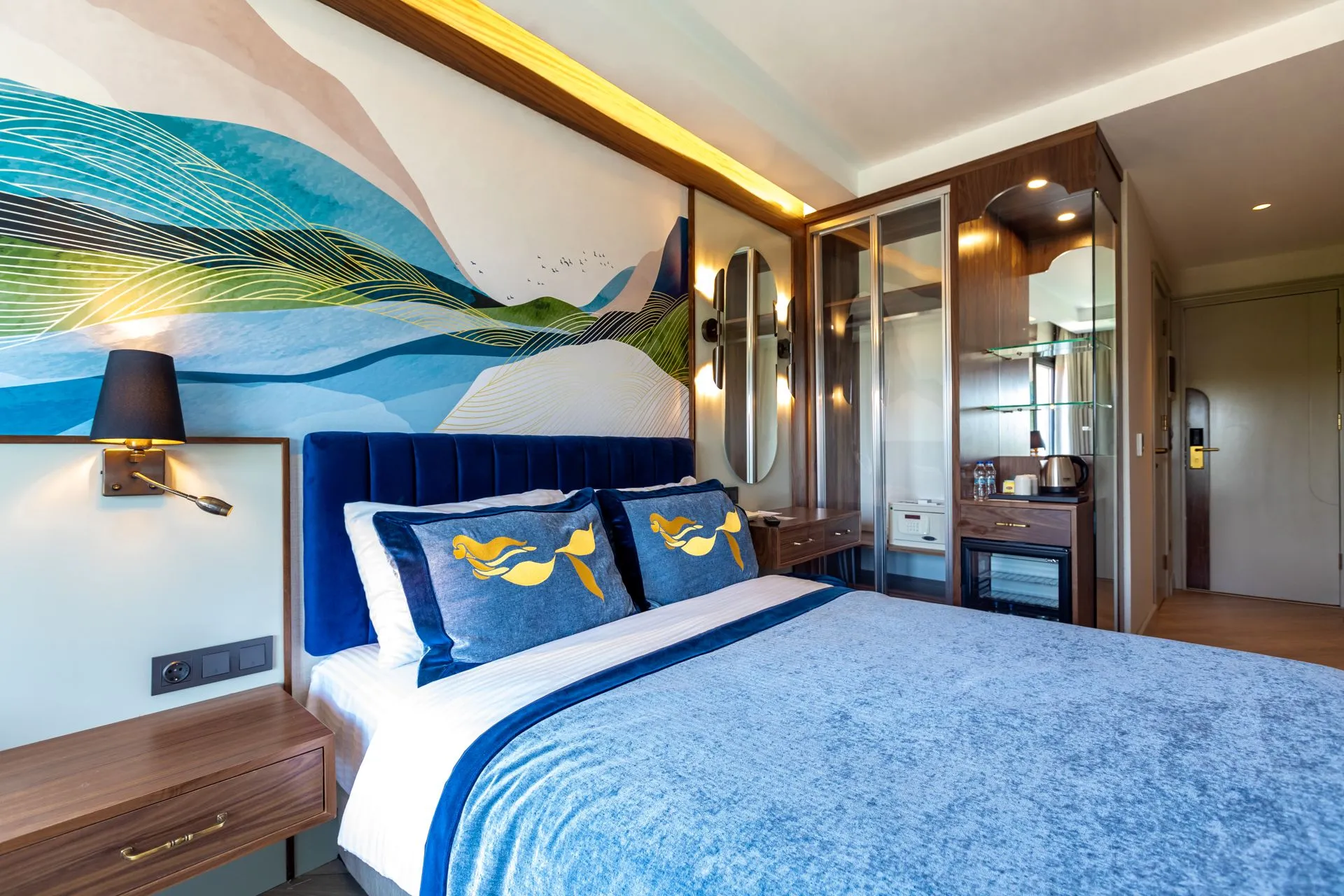 Ein Bett mit marineblauem Samtkopfteil in einem Hotelzimmer, dahinter ein welliges blau-grünes Wandbild und Kissen mit gesticktem Meerjungfrauenschwanz