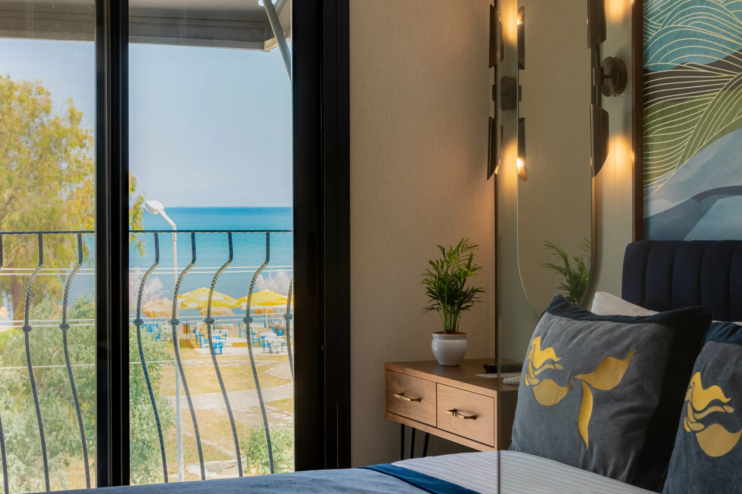 Die Bucht aus dem Zimmerfenster: das Meer hinter dem Balkongeländer und Sonnenschirme am Sand; rechts ein Bett mit Kissen im Meerjungfrau-Motiv