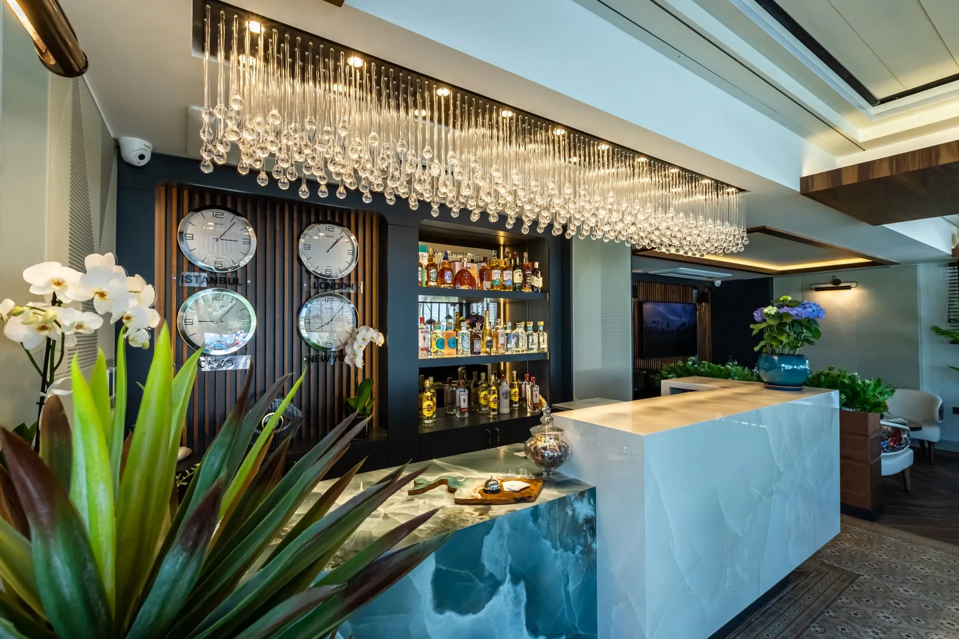 Ein Kristallleuchter in der Hotellobby, Weltuhren an der Wand und eine Rezeption mit hellblauer Onyxtheke