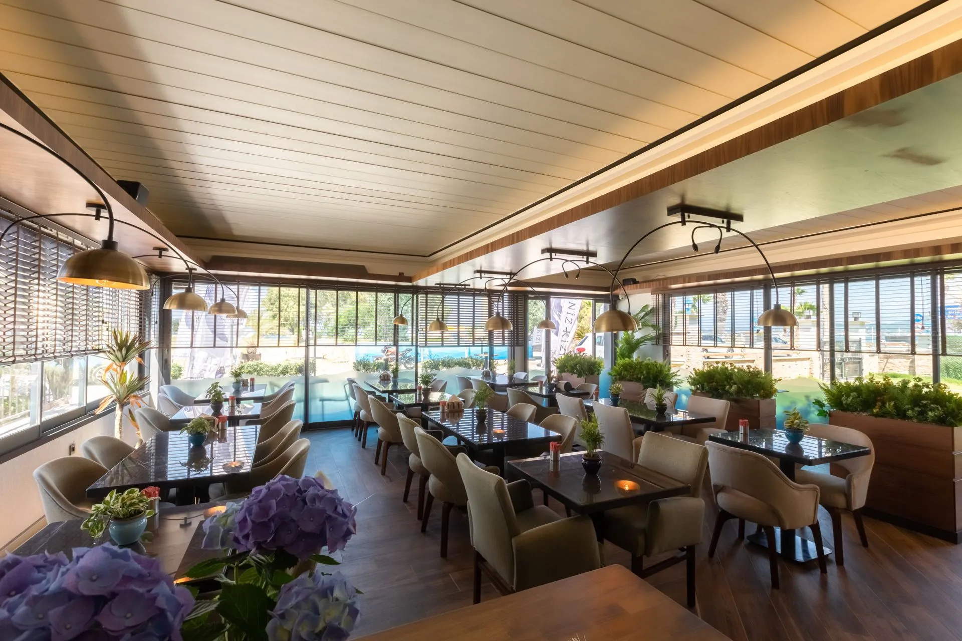 Der Saal des Hotels, der Feiern beherbergt: eine Holzdecke, Messing-Hängelampen, raumhohe Fenster mit Läden, dunkle Holztische und cremefarbene Sessel; vorne lila Hortensien