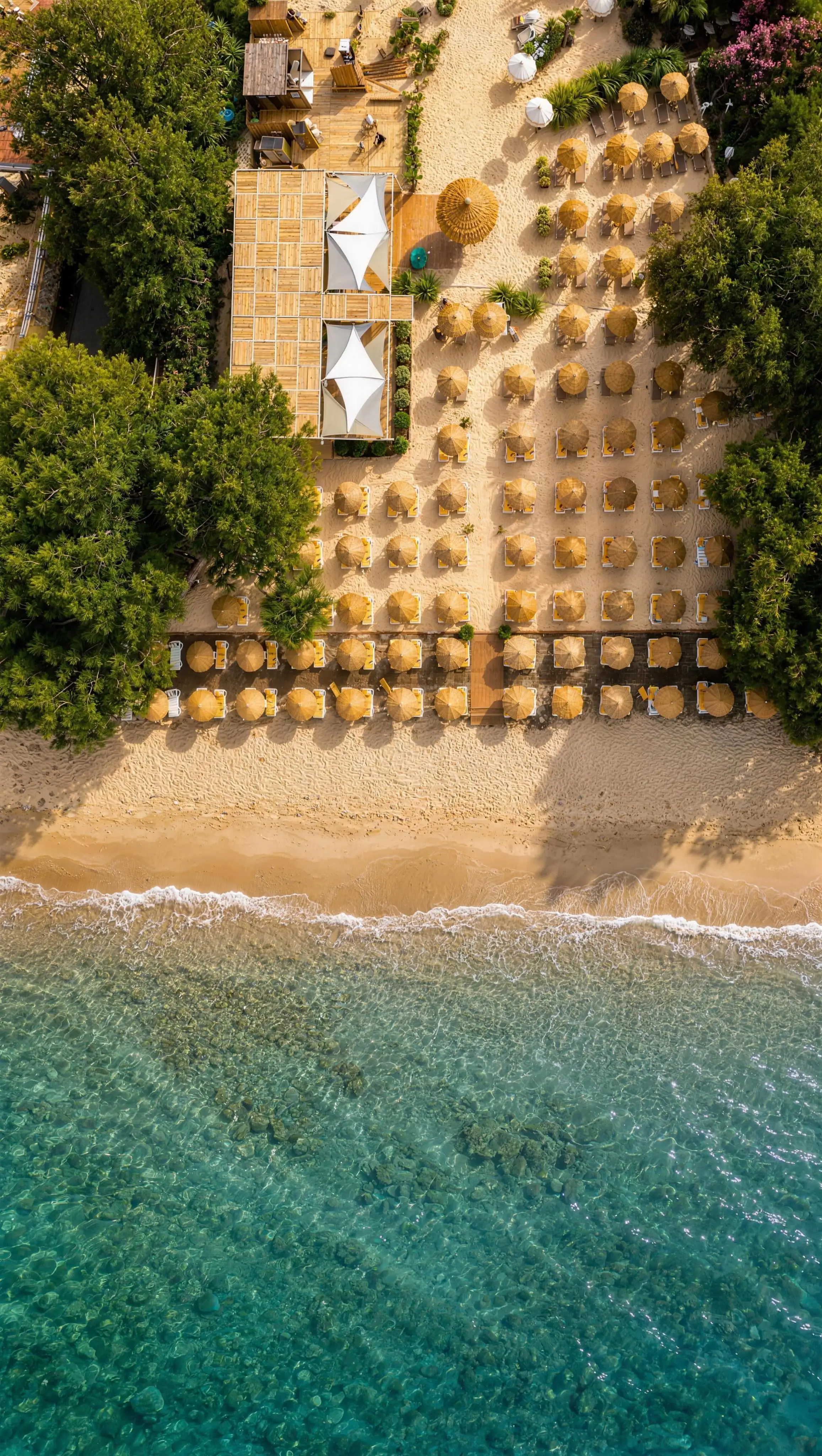 Der Privatstrand von oben: Reihen von Strohschirmen im Sand, eine schäumende Uferlinie und klares türkisfarbenes Wasser