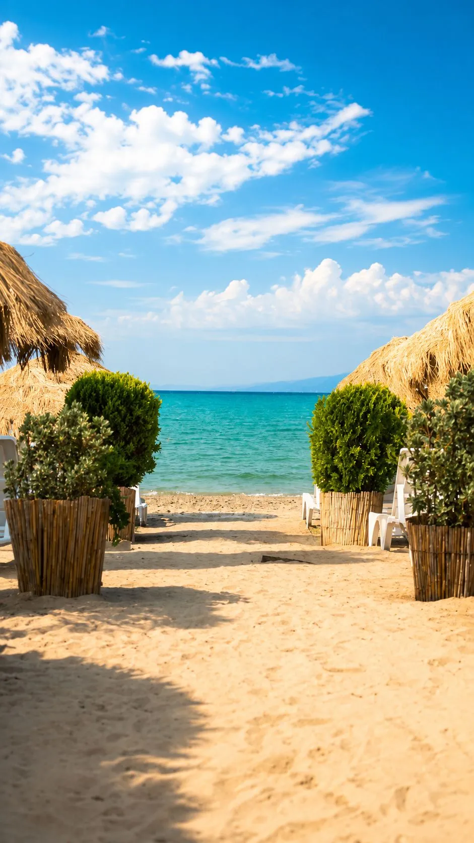 Ein Sandweg vom Hotel hinunter zum Strand: grüne Töpfe beidseitig, Strohschirme und türkisfarbenes Meer am Ende