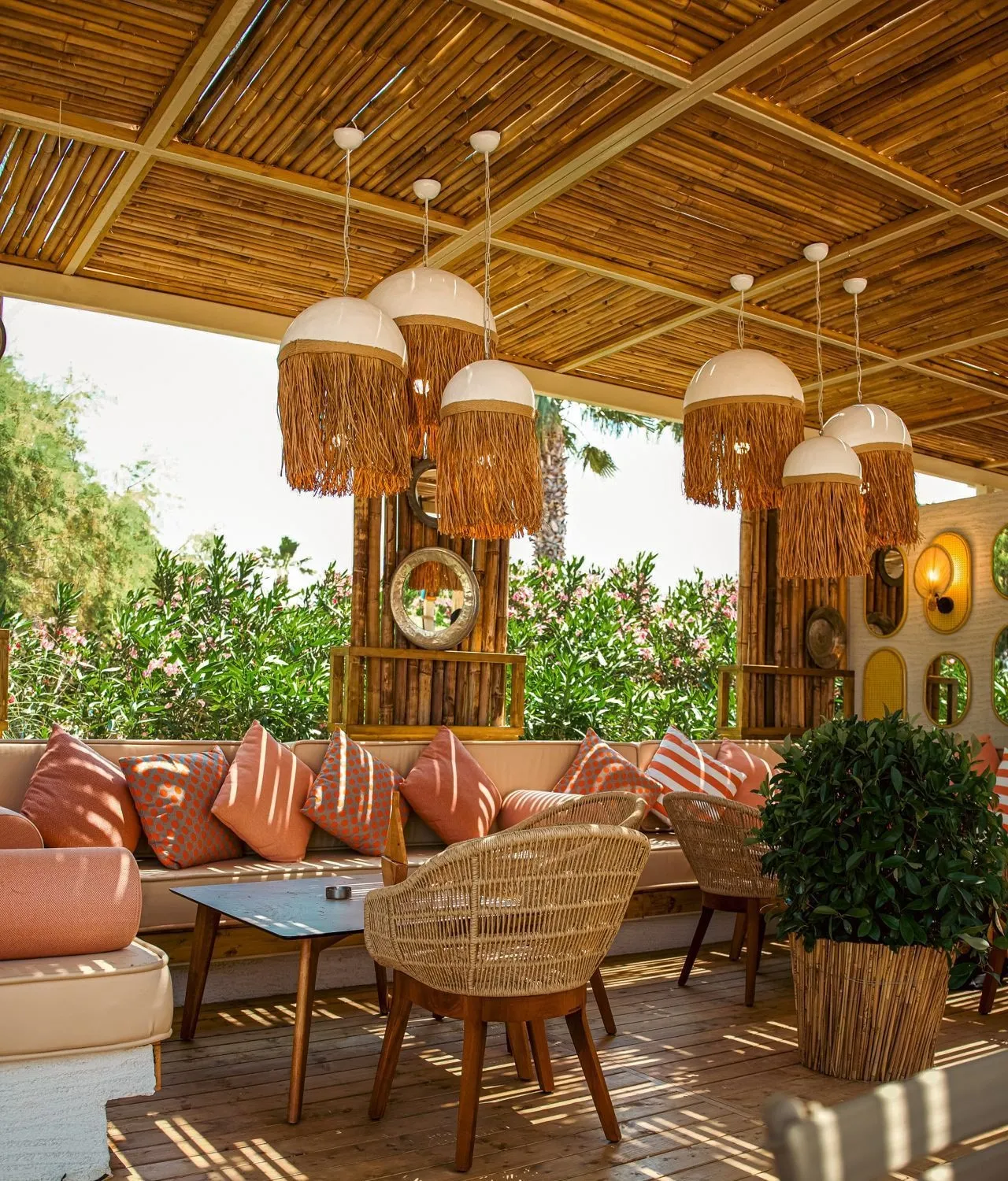 Bastleuchten unter einer Bambuspergola, ein Sofa mit orange-rosa Polstern und Korbstühle; dahinter blühende Sträucher