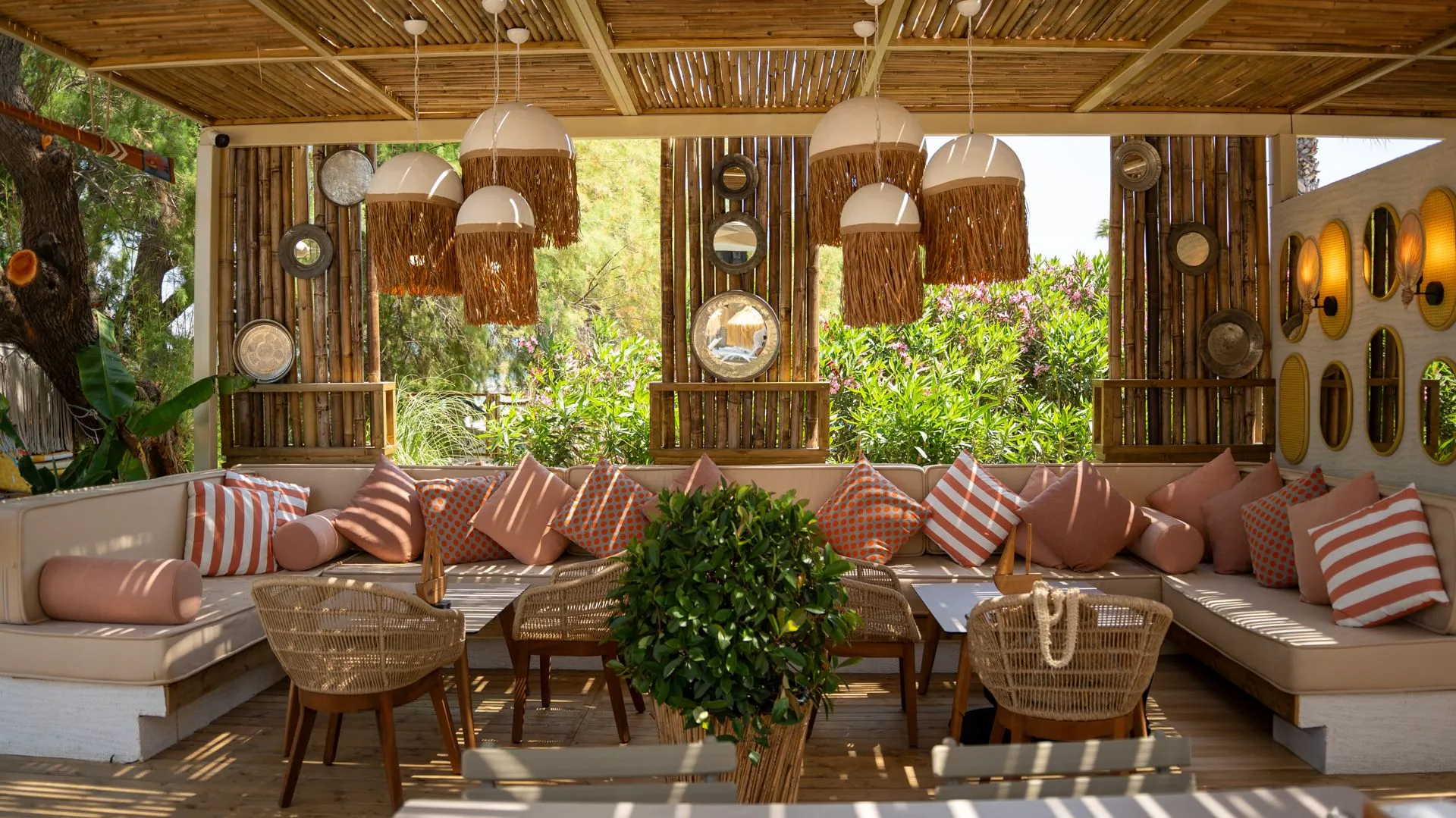 Eine Strandlounge mit Strohdach: eine Sitzgruppe mit rosa und gestreiften Kissen, Rattanstühle und ein grüner Baum in der Mitte