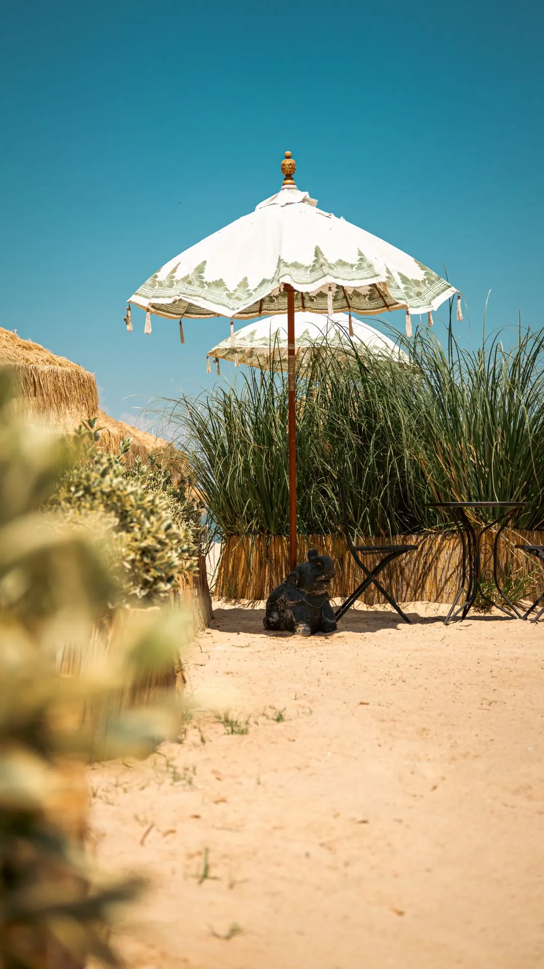 Ein weißer Schirm, geöffnet im Sand, dahinter hohe Gräser und daneben ein kleiner Beistelltisch