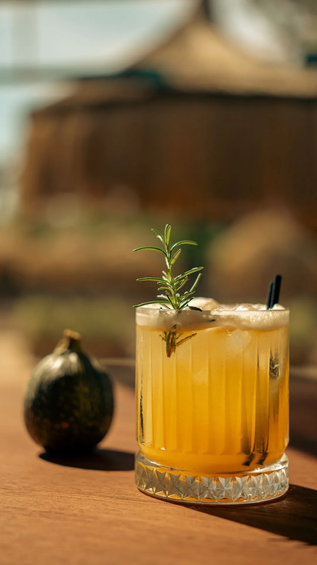 Ein schaumiger gelber Cocktail mit Rosmarin auf einem sonnigen Holztisch; daneben ein kleiner Zierkürbis