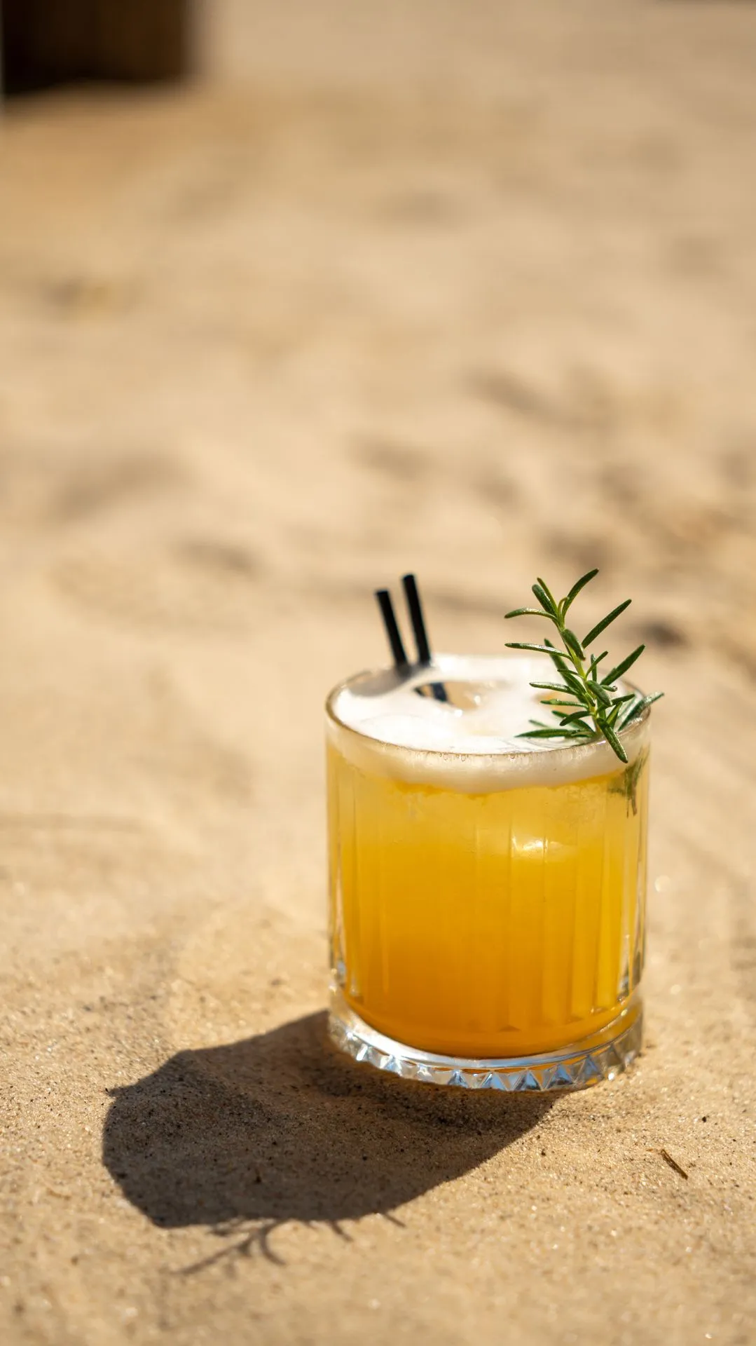 Ein gelber Cocktail, garniert mit Rosmarin, auf dem Sand und sein langer Schatten