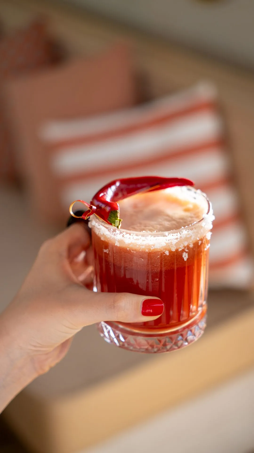 Eine Hand hält einen roten Cocktail mit Salzrand und Chili; dahinter gestreifte Kissen
