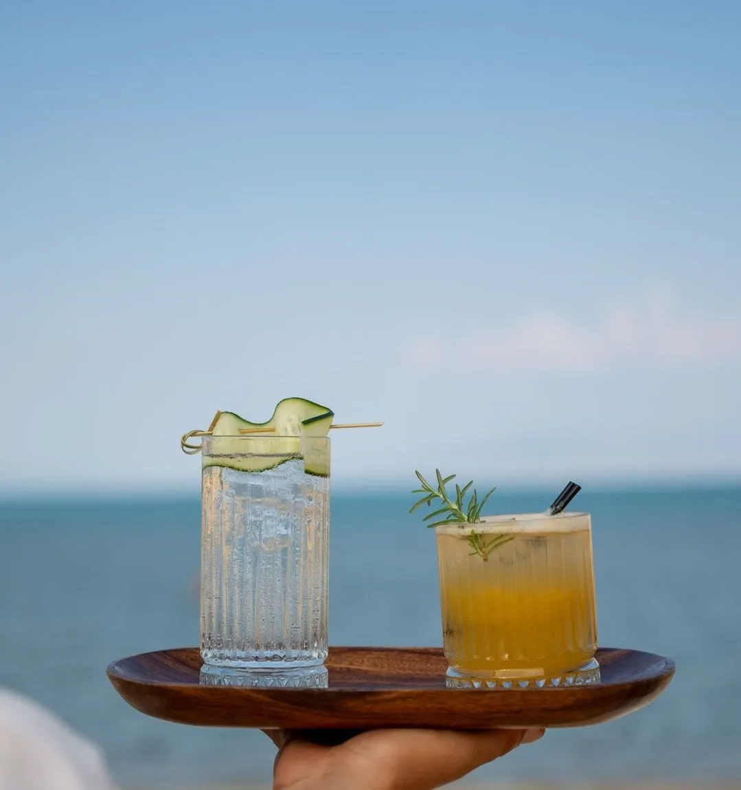 Eine Hand trägt zwei Getränke auf einem Holztablett: einen Longdrink mit Eis und Gurke und einen orangefarbenen Cocktail mit Rosmarin; dahinter das Meer