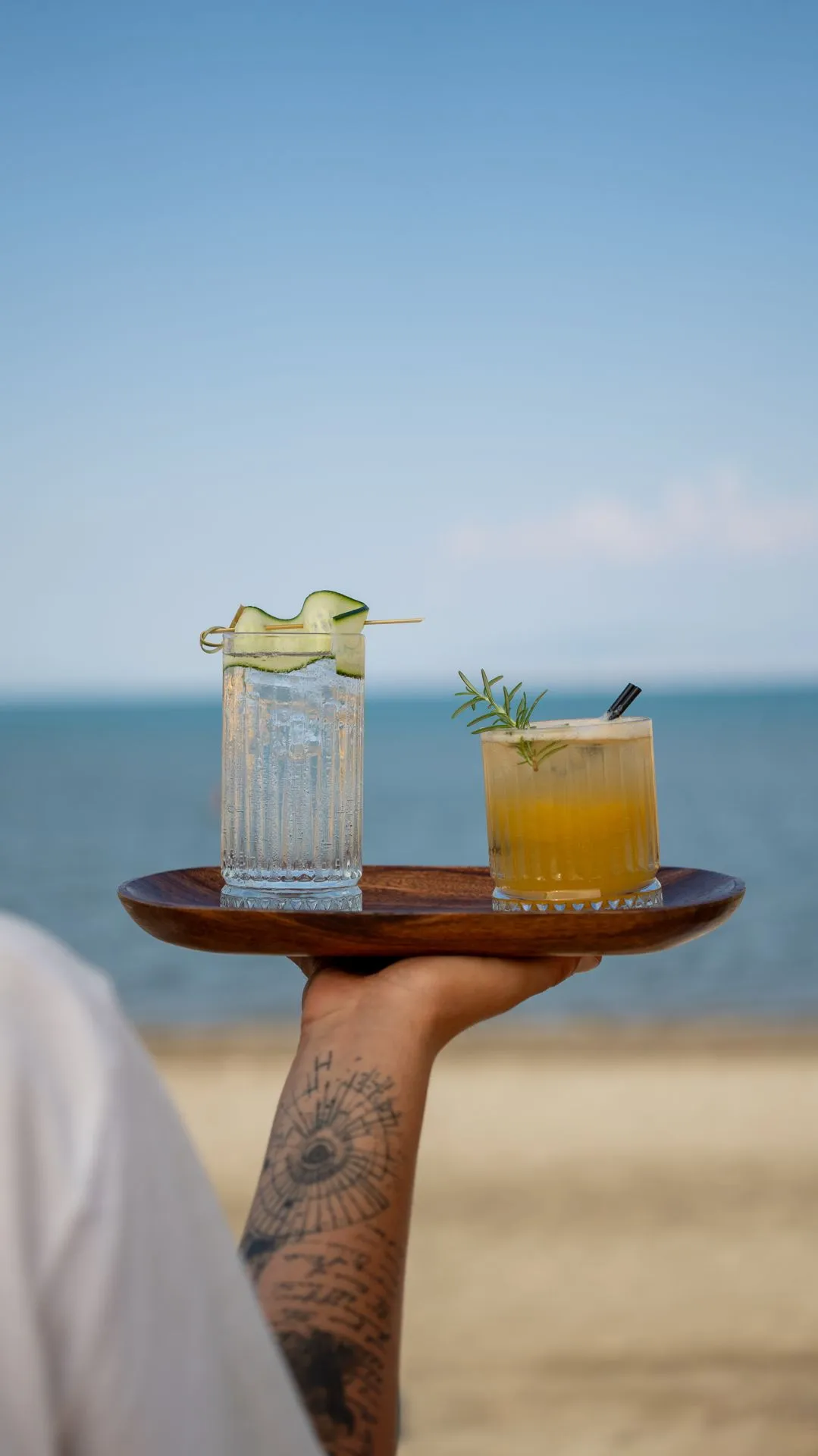 Two refreshing glasses on a wooden tray; the sea horizon of the cove behind