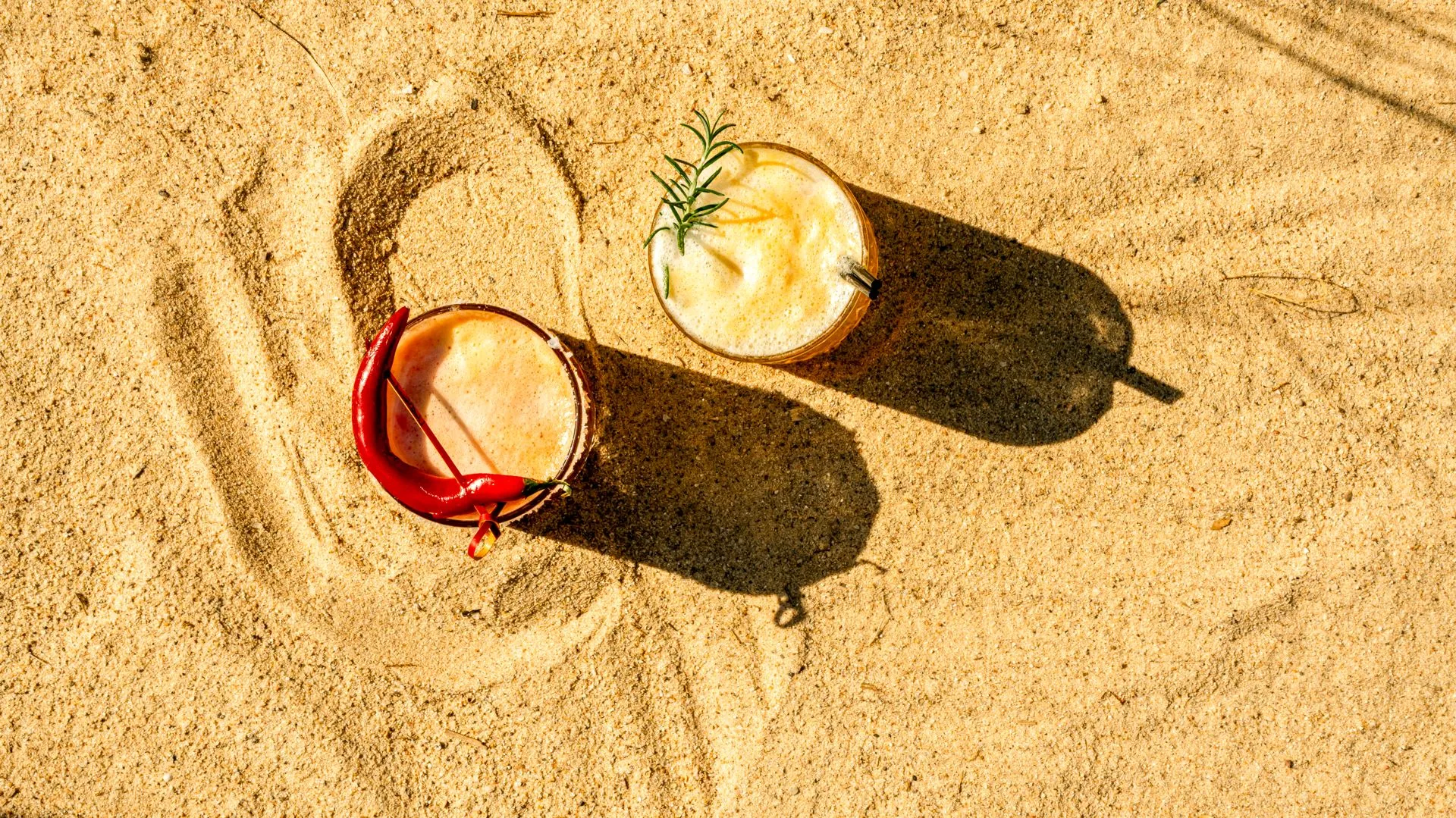 Zwei Cocktails im Sand von oben: ein roter mit Chili und ein gelber mit Rosmarin; in den Sand gezogene Linien und lange Schatten