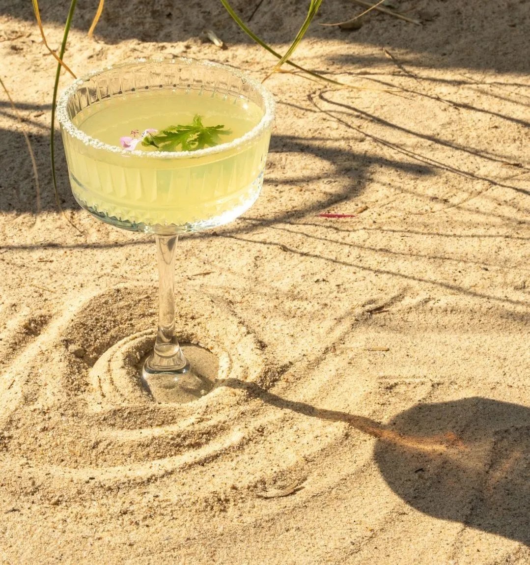 Ein hellgrüner Cocktail mit einer schwimmenden rosa Blüte in einem Stielglas, das im Sand steckt; ringsum in den Sand gezogene Kreise
