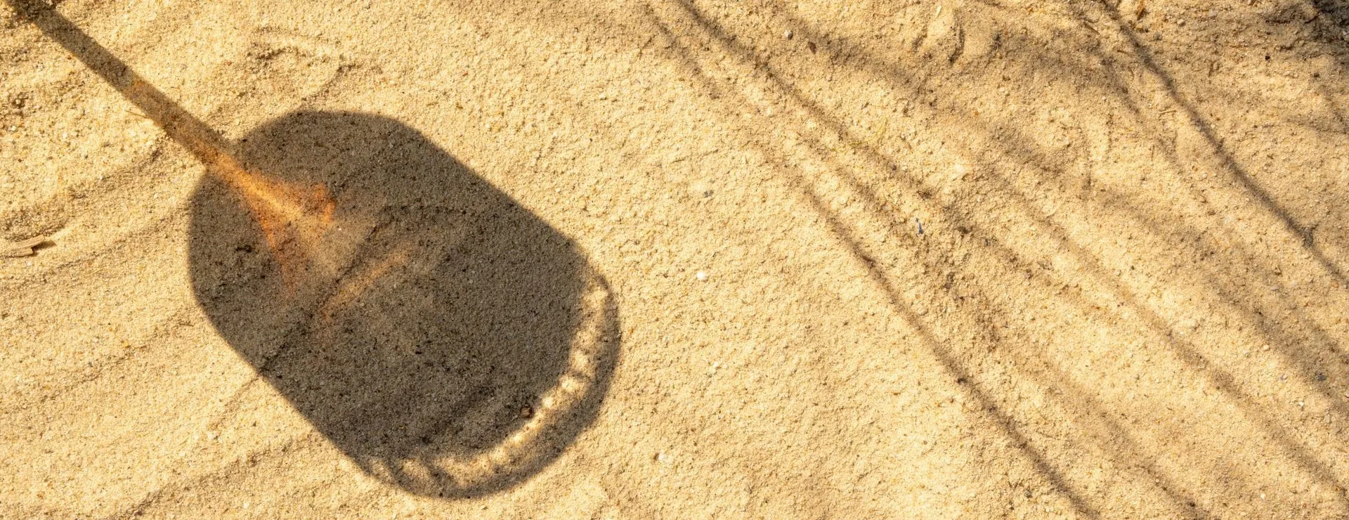 Auf sonnigem Sand ein ovaler Schatten, der wie der eines langstieligen Schlägers oder Paddels aussieht; an seinem Rand orangefarbenes Licht