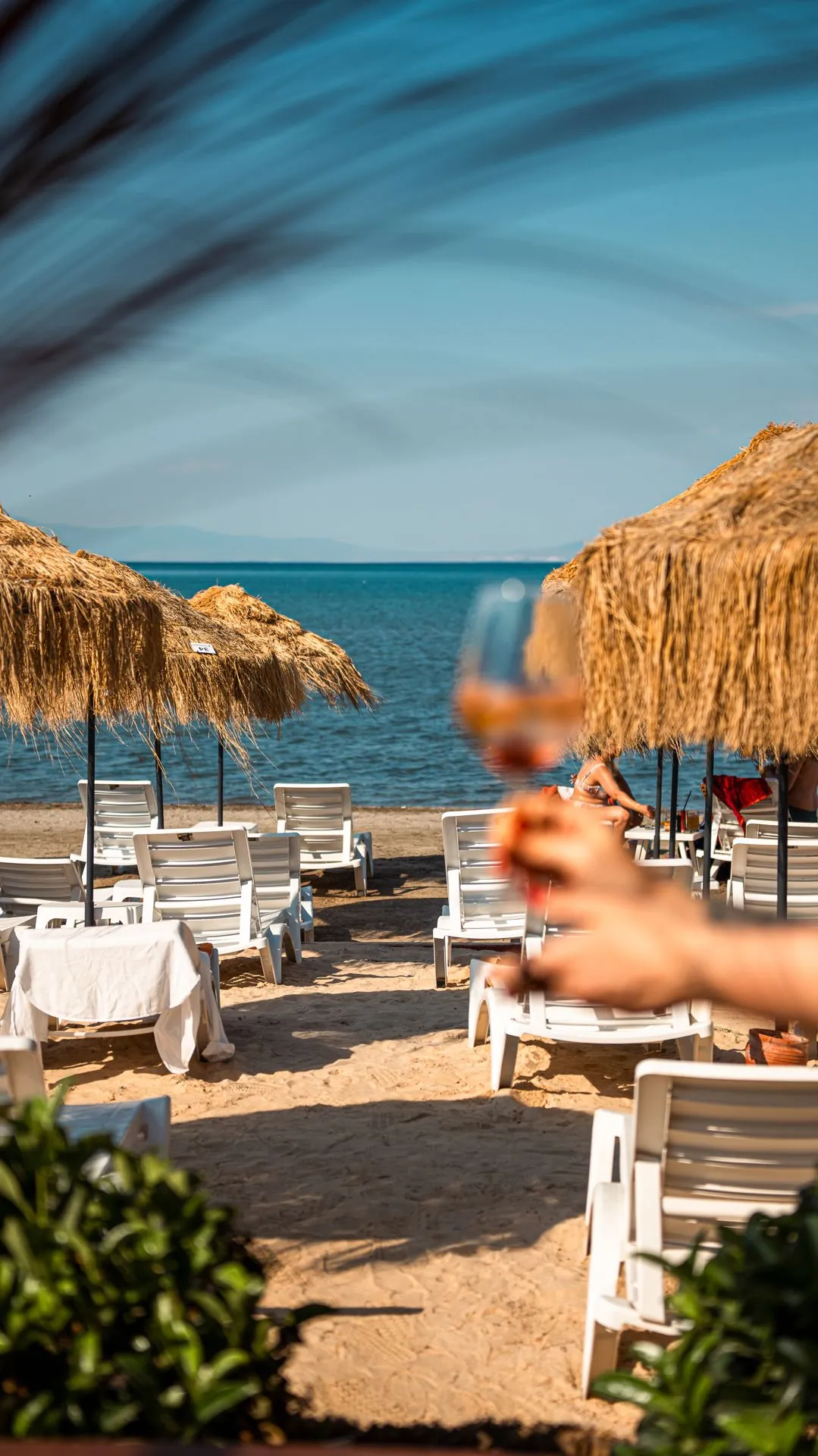 Ein Blick aufs Meer von weiß gedeckten Tischen am Strand: Strohschirme, Liegen und ein Glas im Vordergrund