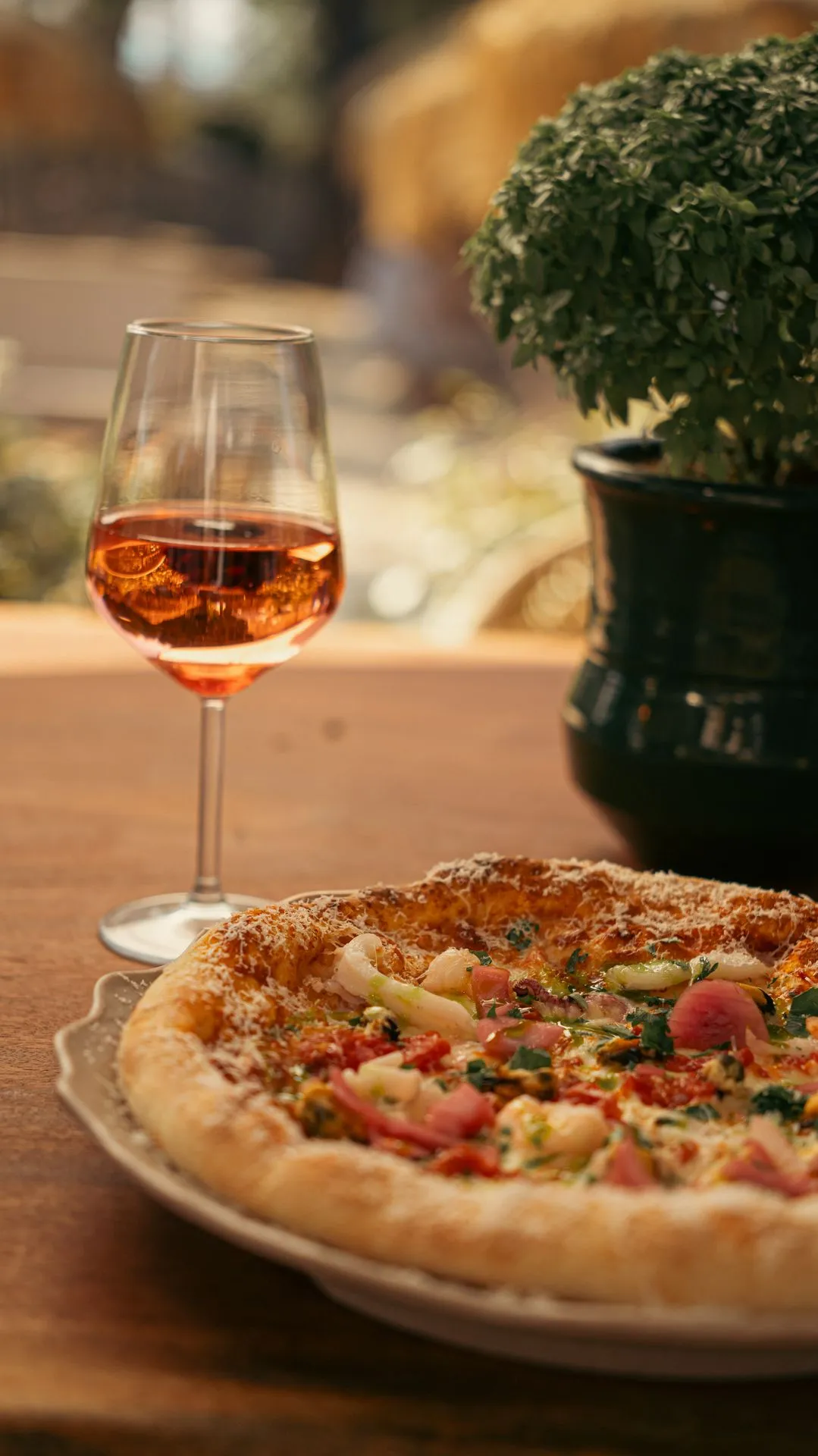 Eine Pizza mit Meeresfrüchten auf einem Holztisch, daneben ein Glas Rosé und eine grüne Topfpflanze