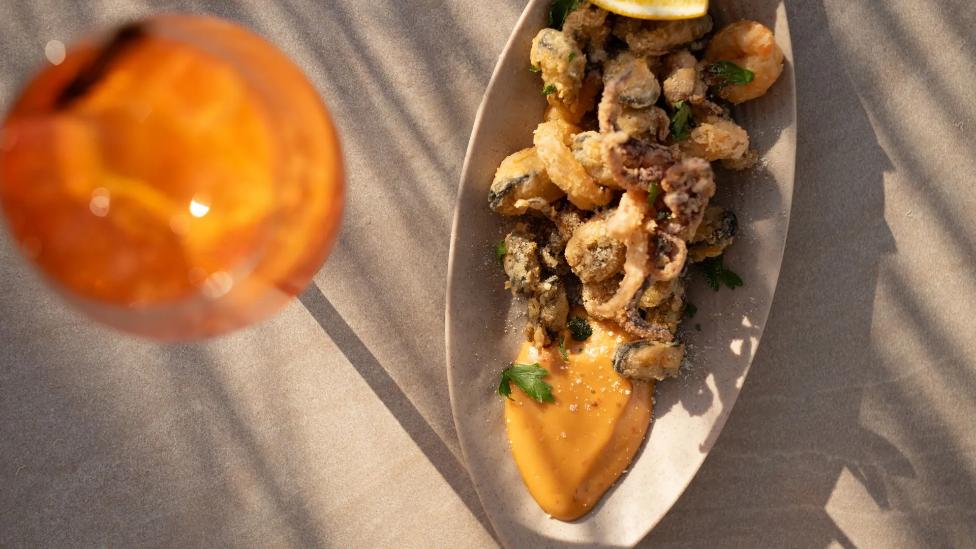 Frittierte Calamari, Muscheln und Garnelen auf einem kahnförmigen Keramikteller, am Ende orange Sauce und Petersilie; links ein unscharfer Cocktail und Palmenschatten auf dem Tuch
