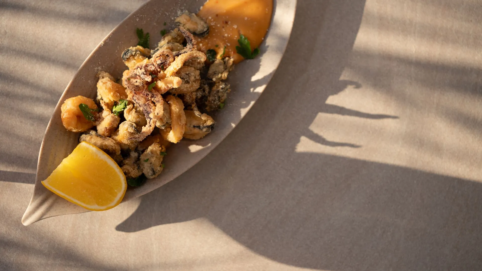 Fried seafood on a handmade ceramic boat-shaped plate with a lemon wedge and an orange sauce; palm-frond shadows across a sand-coloured surface