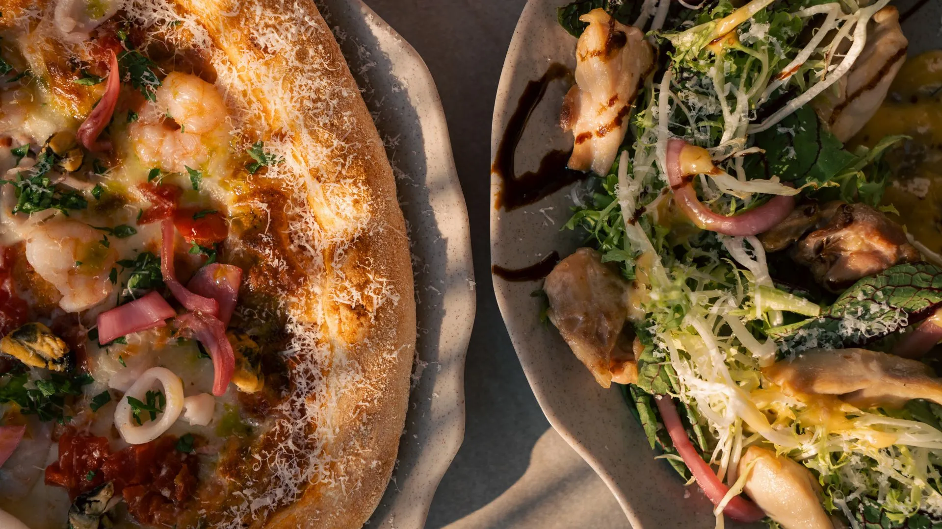 Two plates side by side on the same table: on the left a pizza with prawns and calamari rings; on the right a leafy salad with red onion, grated cheese and balsamic
