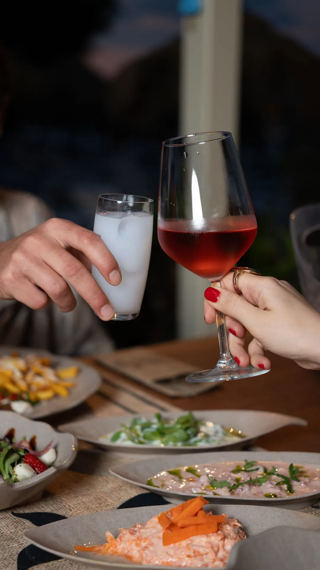 Two hands clinking glasses at a table in evening light; meze plates in front of them