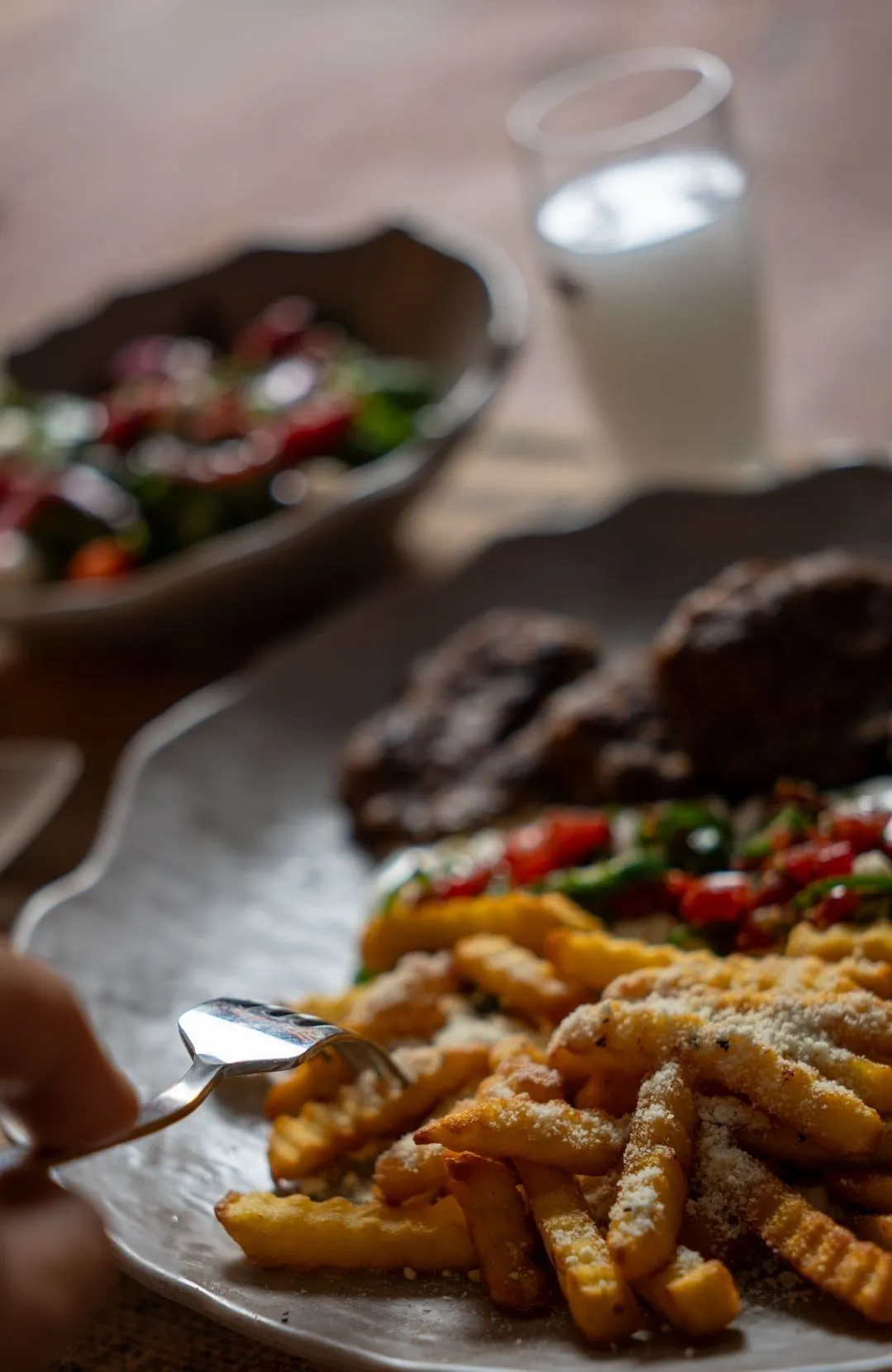 Wellenpommes mit geriebenem Käse; dahinter Köfte und eine Tomaten-Paprika-Beilage, weiter hinten eine Salatschale und ein weißes Getränk; von links greift eine Gabel zu