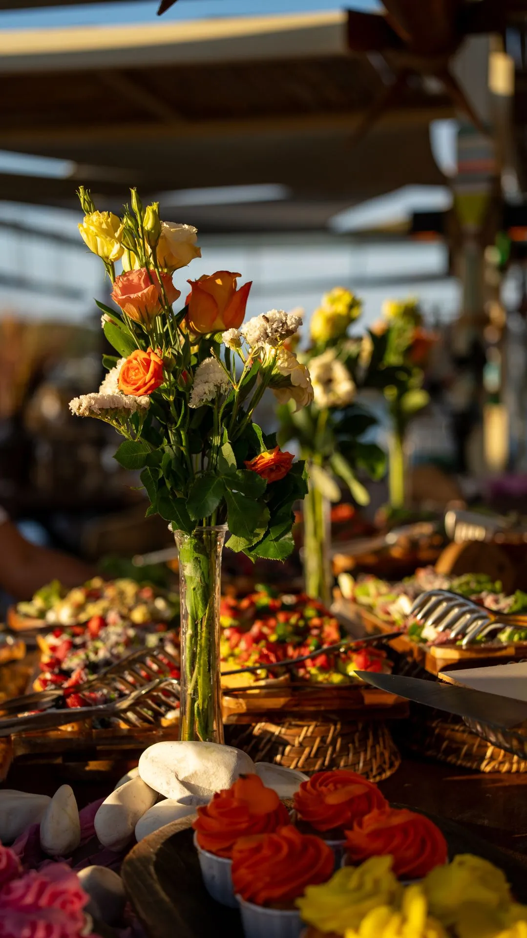 Ein Tisch im Abendlicht: orangefarbene Rosen in einer Glasvase, dahinter Mezze-Tabletts und Holzteller
