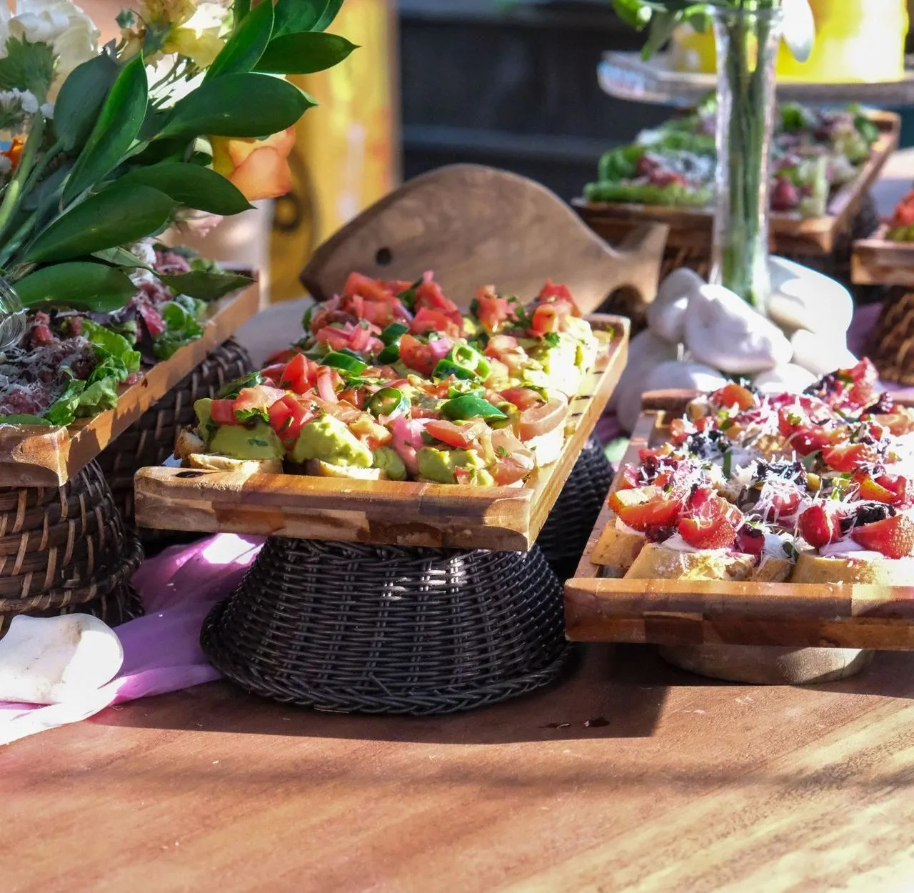 Ein Buffettisch: geröstetes Brot mit Avocado und Tomate auf Holzbrettern auf Weidenkörben, daneben Brote mit roten Früchten und Blumen