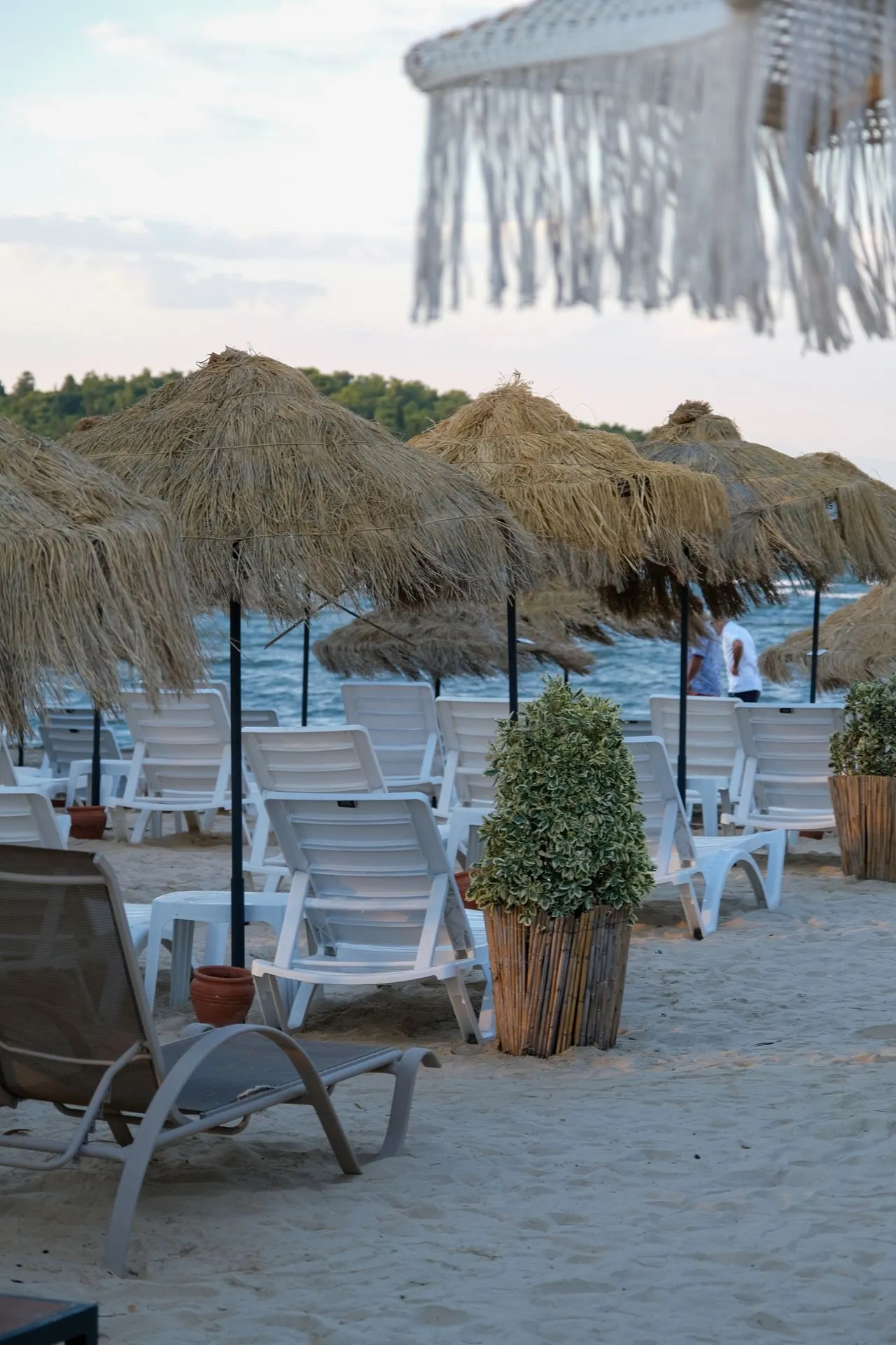 Der Strand auf Augenhöhe: Reihen weißer Liegen, Strohschirme und Sträucher in Töpfen; dahinter das Meer und ein bewaldetes Ufer