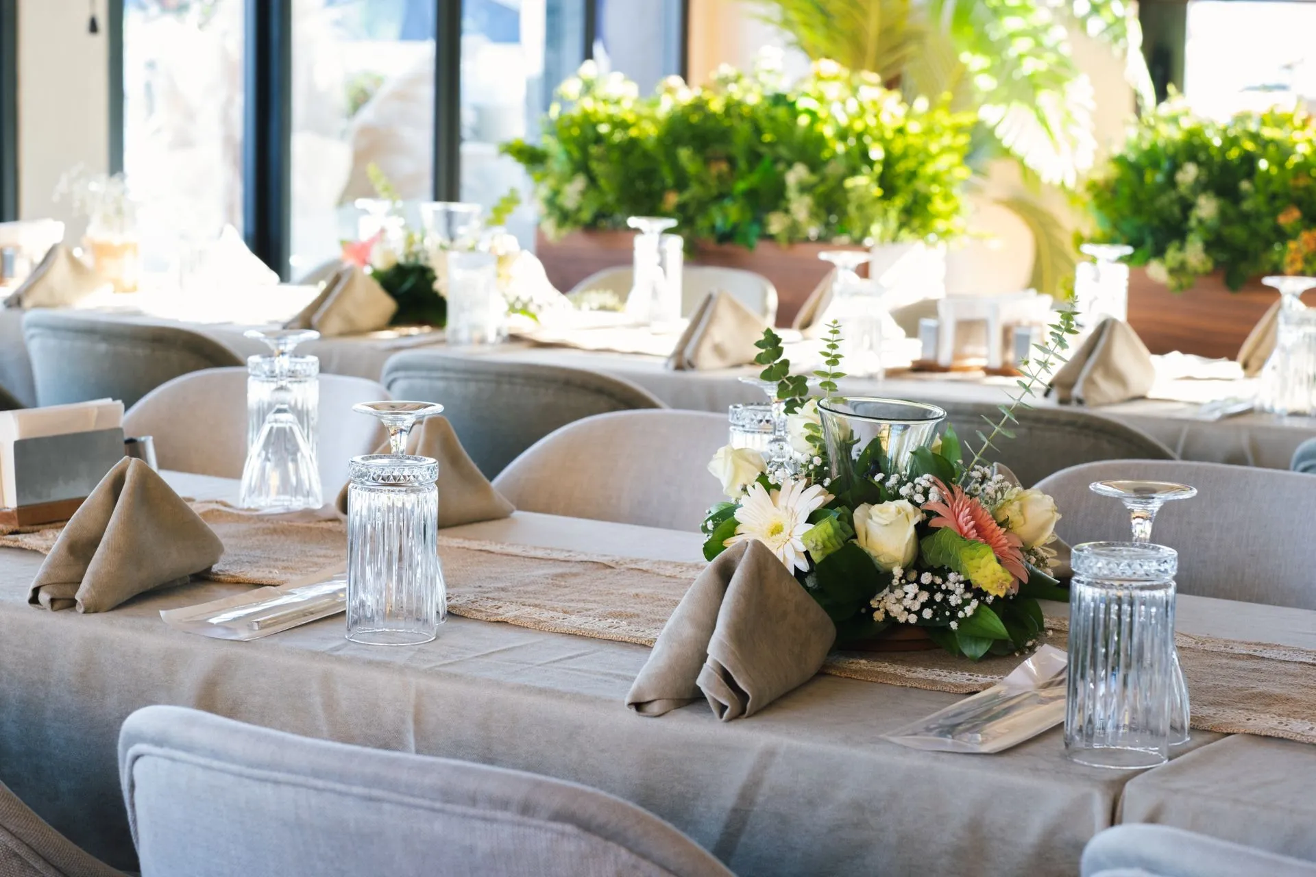 Ein langer, für eine Feier gedeckter Tisch: gefaltete Leinenservietten, umgedrehte Gläser, Gerbera- und Rosengestecke, Topfgrün und sonnendurchflutete Fenster dahinter