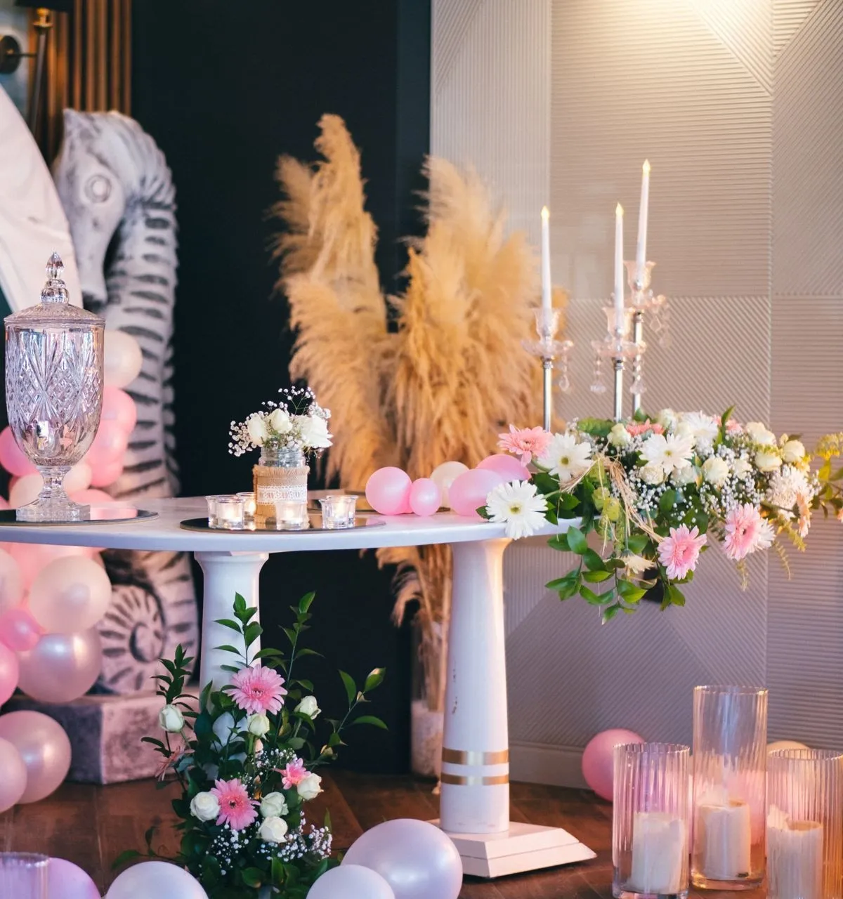 Eine Feierecke: Blumen und Kerzen auf einem weißen Tisch, gläserne Kerzenständer, Pampasgras und daneben eine Seepferdchenskulptur; rosa und weiße Luftballons am Boden