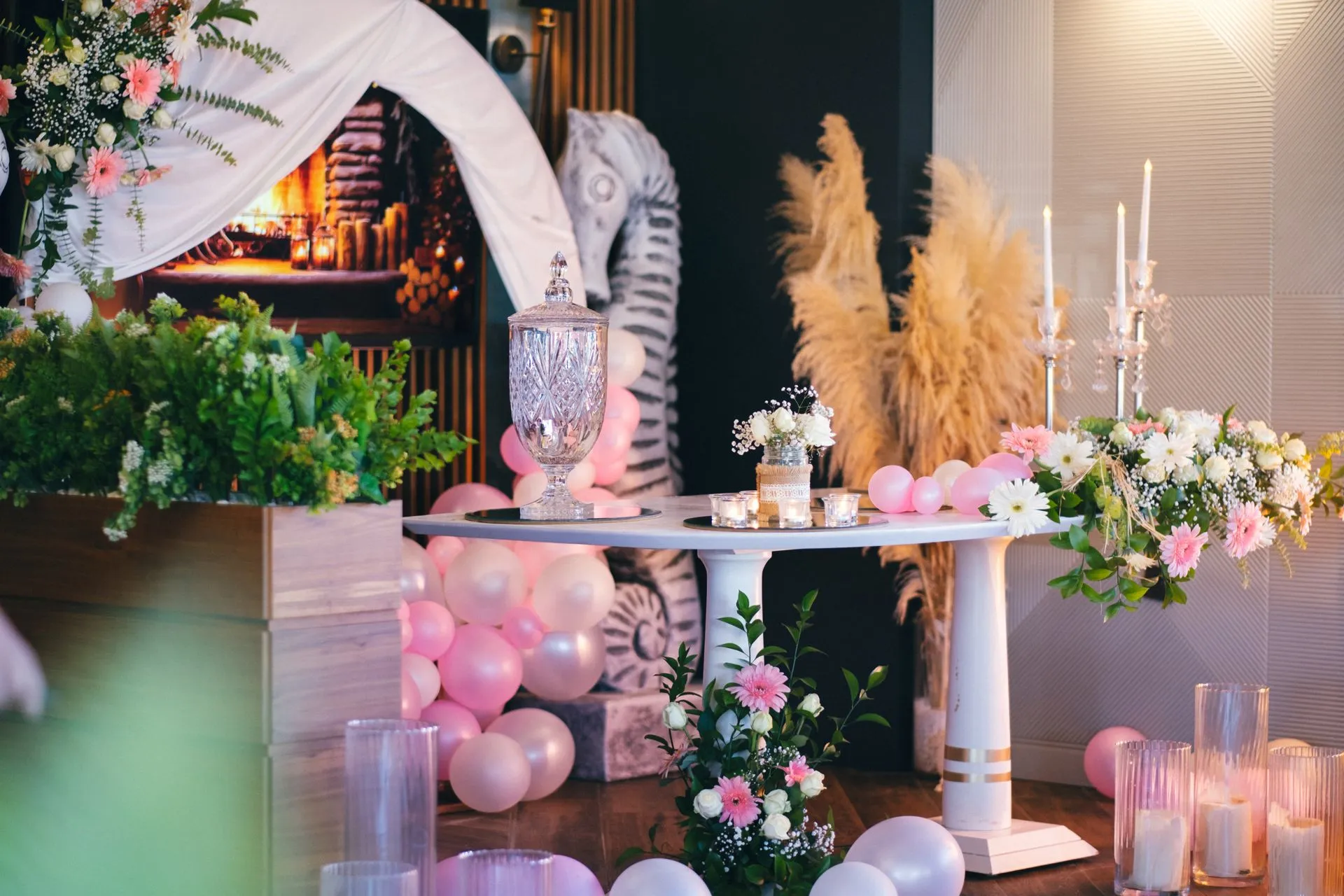 An indoor celebration setup: candles and a crystal jar on a round white table, surrounded by pink balloons, pampas grass and floral arrangements; a seahorse relief behind