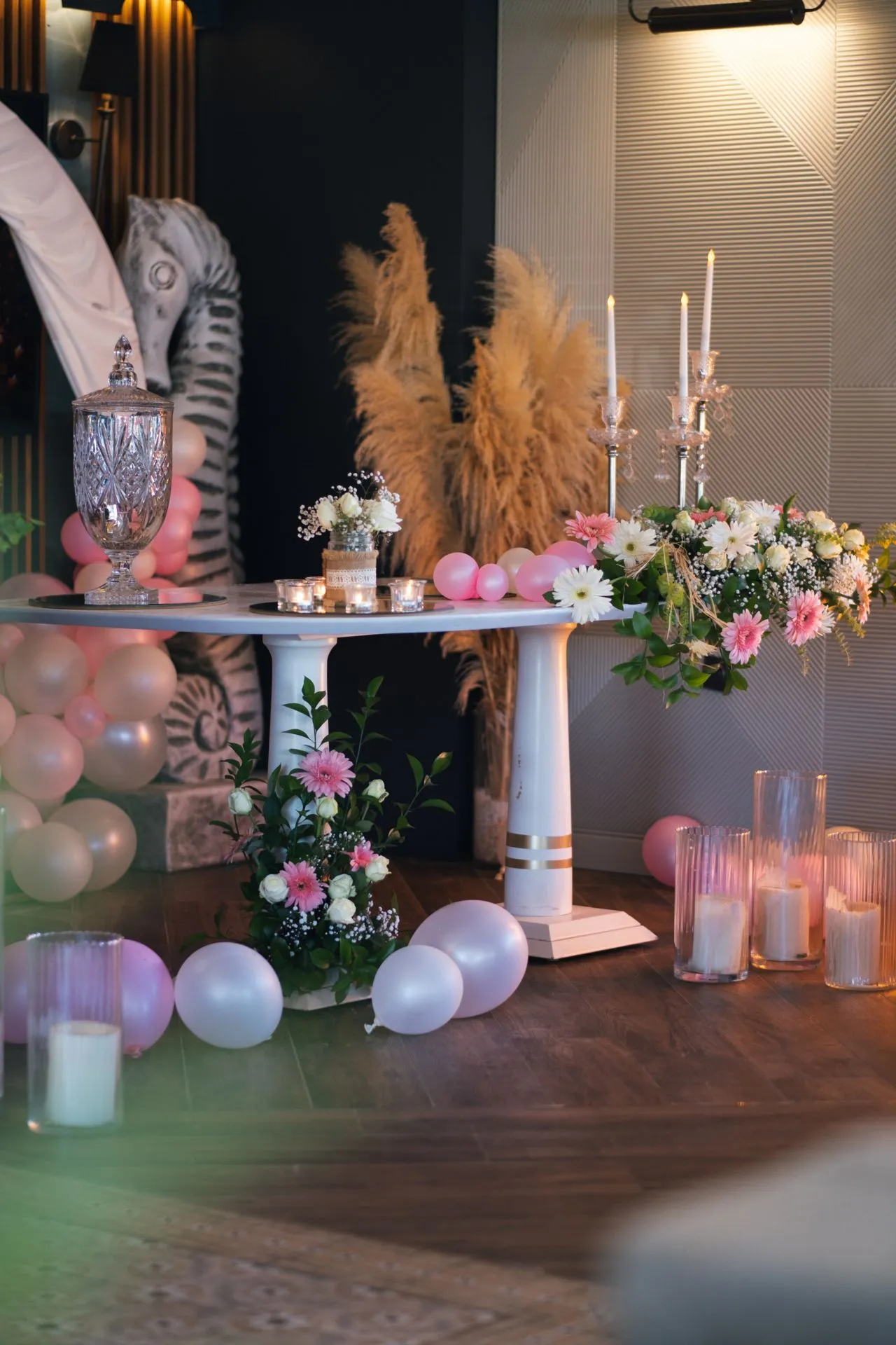 Ein für eine Verlobung vorbereiteter Aufbau: ein Kristall-Getränkespender auf einem weißen Tisch, Teelichter darum herum, rosa und cremefarbene Ballons, Pampasgras und Blumengestecke