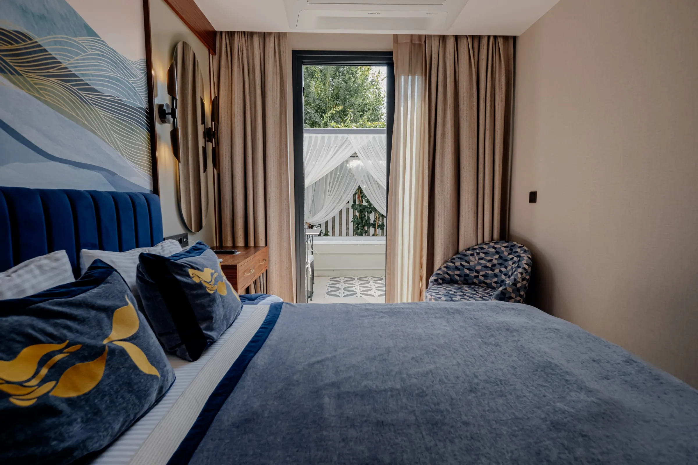 Wellen-Wandbild und ovaler Spiegel hinter dem Bett; hinter der Tür die von Voile beschattete Terrasse und ihr gemusterter Boden